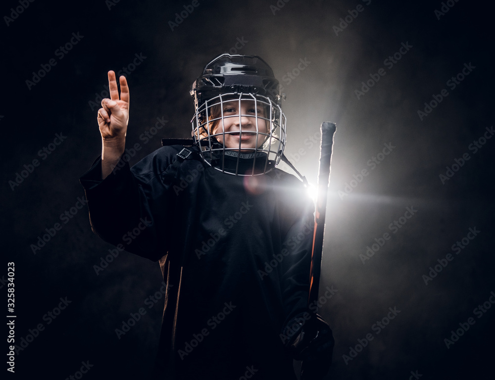 Young blonde sporty boy, ice hockey player, posing in a dark studio for a photoshoot, wearing an ice-skating uniform, helmet, hockey stick, showing a victory gesture with his hand and smiling