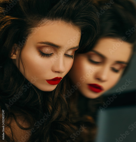 Model lies on mirror eyes closed. Close-up portrait princess. Bright evening make up red lips brown shine glitter shadows. Elegant wavy hairstyle long healthy hair brunette. face mirroring backdrop