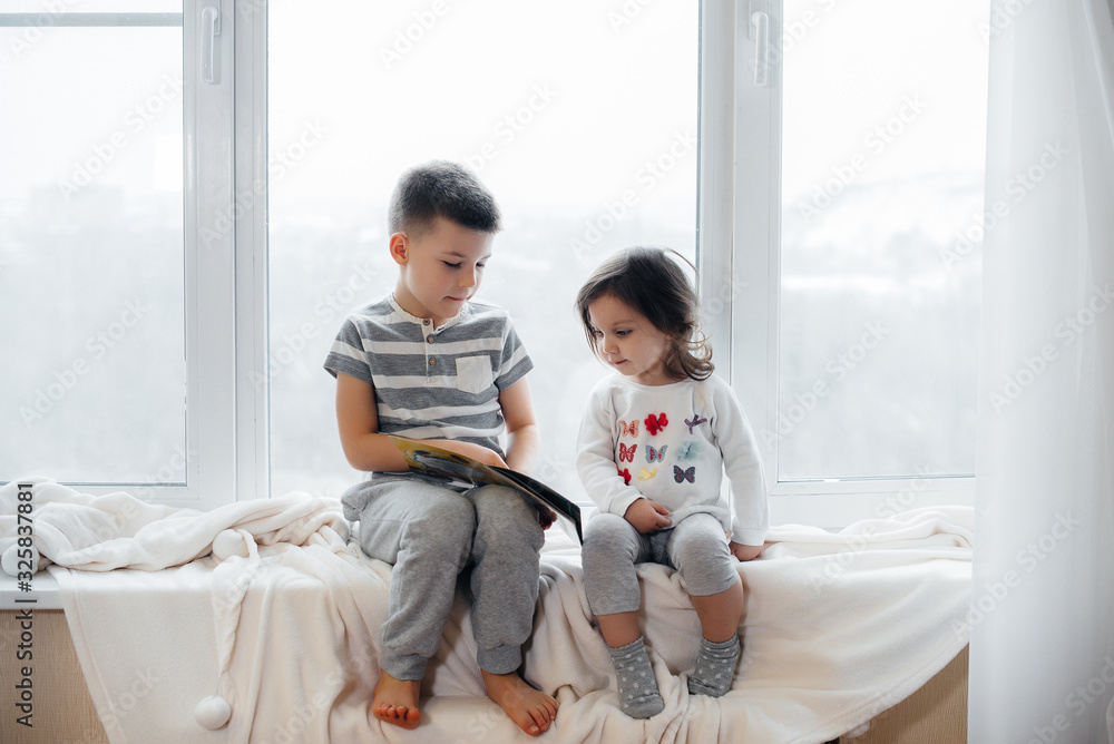 Brother and sister are sitting on the windowsill and reading a book. Happiness, family Brother and sister are sitting on the windowsill and reading a book. Happiness, family