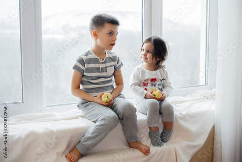 Brother and sister are sitting on the windowsill playing and eating apples. Happiness