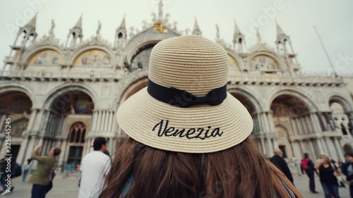 Brunette girl in hat stands on San Marco Square, looks to Basilica San Marco
