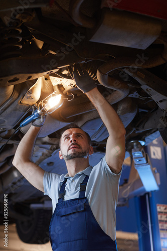 Low angle portrait of mature mechanic looking under car on lift and holding lamp light during inspection in auto repair workshop, copy space