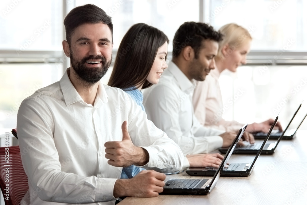 Obraz premium Smiling cheerful caucasian bearded man with thumb up in office. Young adult man with beard smiling with manager, accompanied with colleagues and shows thumb up.