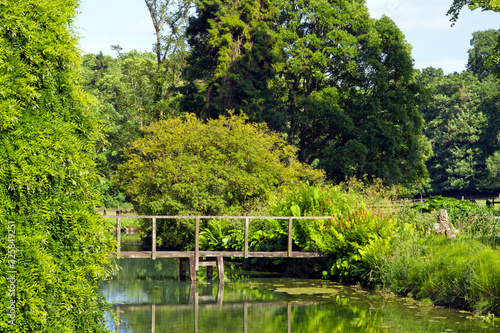 Old wooden bridge across small river with lush vegetation growing on the banks, in an English countryside on a summer sunny day .