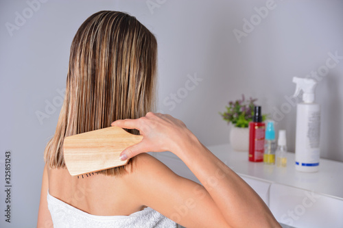 Woman with comb brushing her wet blonde hair after showering at home. Cares about healthy and clean hair. Beauty salon concept. Girl wears white towel around chest. Close up, selective focus