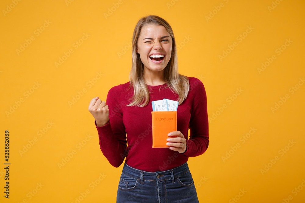 Laughing young blonde woman girl in casual clothes posing isolated on yellow orange wall background. People lifestyle concept. Mock up copy space. Hold passport tickets, doing winner gesture blinking. Laughing young blonde woman girl in casual clothes posing isolated on yellow orange wall background. People lifestyle concept. Mock up copy space. Hold passport tickets, doing winner gesture blinking.