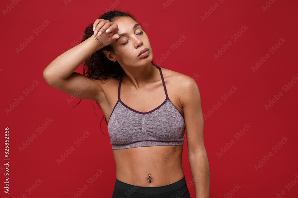 Obraz premium Tired young african american sports fitness woman in sportswear posing working out isolated on red wall background. Sport exercises healthy lifestyle concept. Putting hand on head keeping eyes closed.