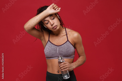 Tired african american fitness woman in sportswear isolated on red background. Sport exercises healthy lifestyle concept. Hold water bottle, working out, listen music with earpods, put hand on head.