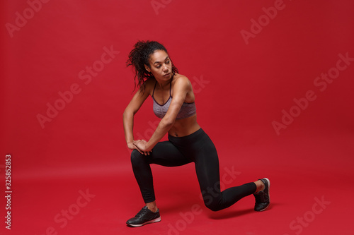 Attractive young african american sports fitness woman in sportswear posing working out isolated on red wall background. Sport exercises healthy lifestyle concept. Stretching her legs, doing lunges.