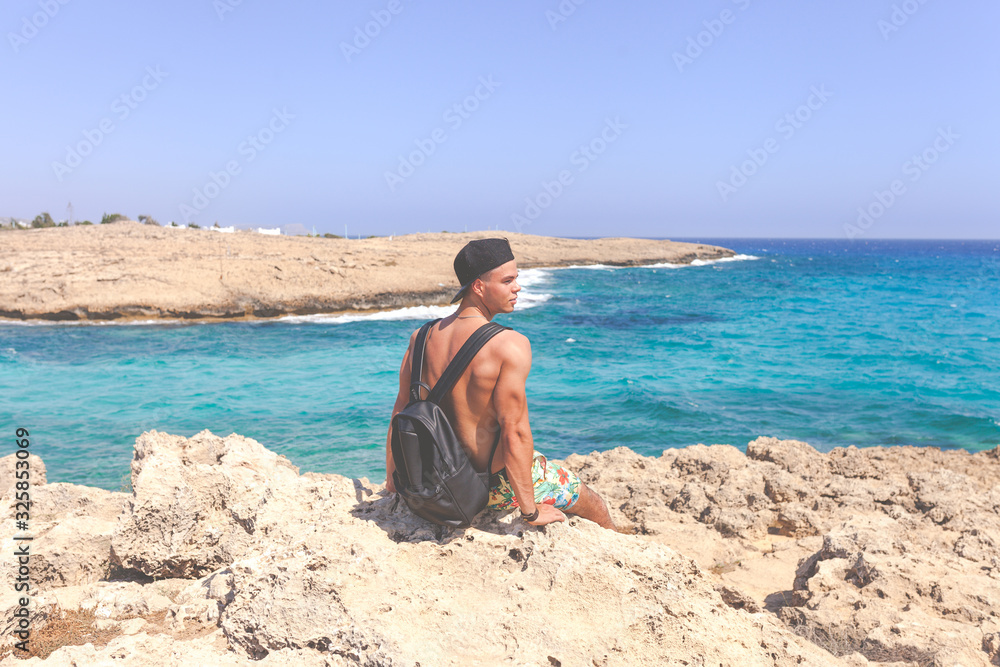 Athletic young man in cap sitting around Natural Rock Bridge at Cape Greco near Ayia Napa in the evening light. Cyprus. Healthy lifestyle Athletic young man in cap sitting around Natural Rock Bridge at Cape Greco near Ayia Napa in the evening light. Cyprus. Healthy lifestyle