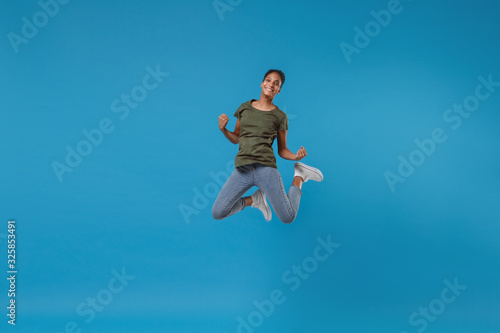 Joyful young african american woman girl in casual clothes posing isolated on bright blue wall background studio portrait. People lifestyle concept. Mock up copy space. Jumping, doing winner gesture.