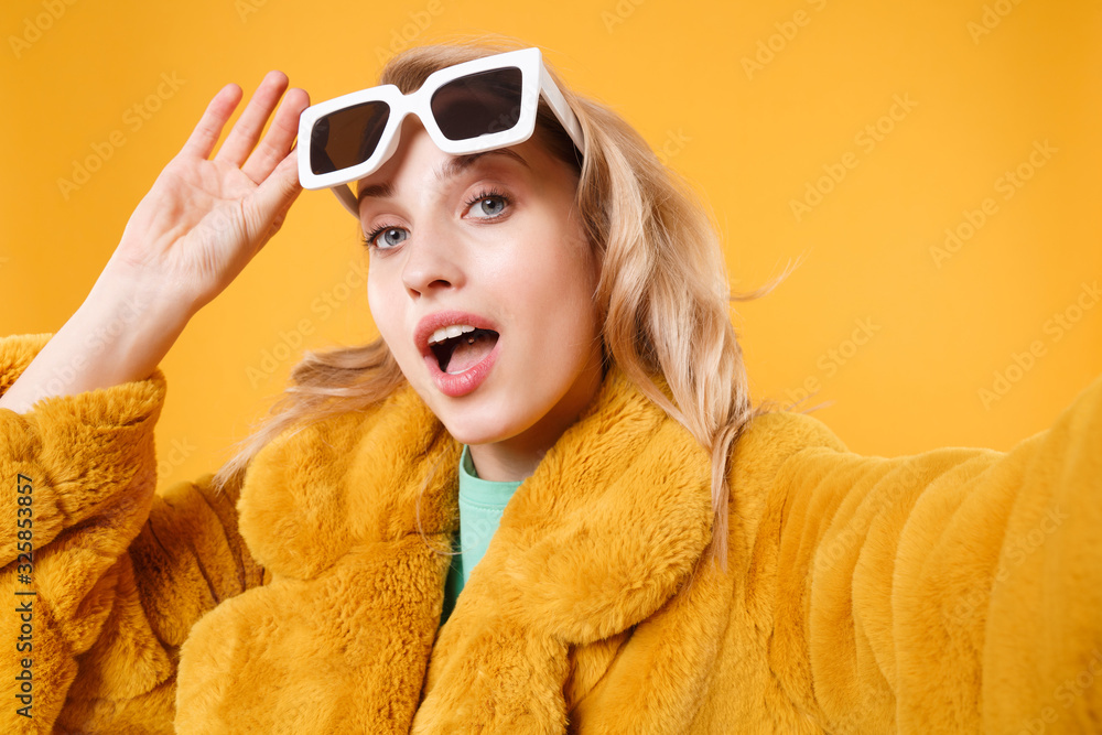 Close up of stunning young blonde woman in yellow fur coat, dark sunglasses posing isolated on orange wall background. People lifestyle concept. Mock up copy space. Doing selfie shot on mobile phone.