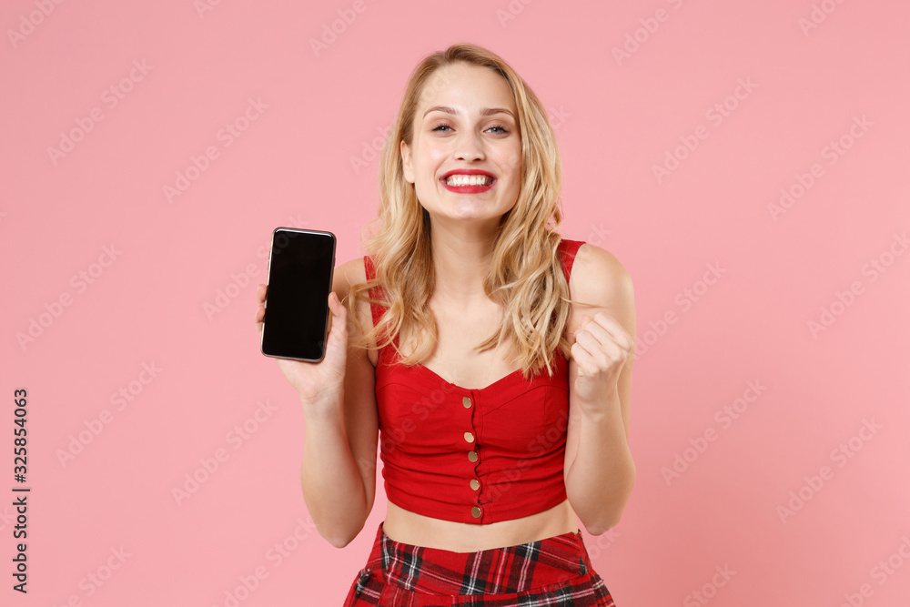 Smiling young blonde woman in red sexy clothes posing isolated on pastel pink background. People lifestyle concept. Mock up copy space. Hold mobile phone with blank empty screen doing winner gesture. Smiling young blonde woman in red sexy clothes posing isolated on pastel pink background. People lifestyle concept. Mock up copy space. Hold mobile phone with blank empty screen doing winner gesture.