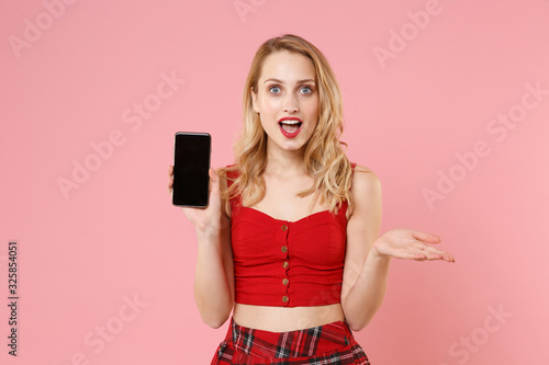 Excited young blonde woman girl in red sexy clothes posing isolated on pastel pink background. People lifestyle concept. Mock up copy space. Hold mobile phone with blank empty screen spreading hands.