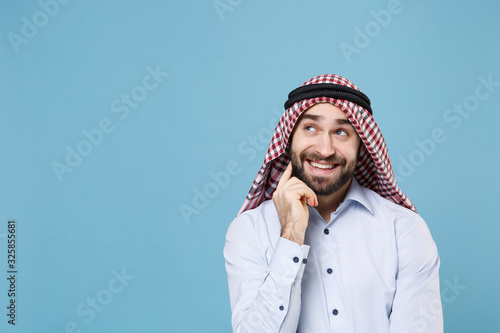 Pensive young arabian muslim man in keffiyeh kafiya ring igal agal casual clothes isolated on pastel blue background. People religious lifestyle concept. Mock up copy space. Put hand prop up on chin. photo