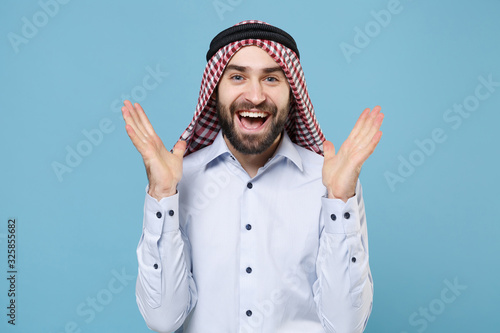 Excited bearded young arabian muslim man in keffiyeh kafiya ring igal agal casual clothes isolated on pastel blue background. People religious lifestyle concept. Mock up copy space. Spreading hands. photo