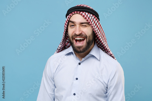 Cheerful bearded young arabian muslim man in keffiyeh kafiya ring igal agal casual clothes posing isolated on pastel blue background. People religious lifestyle concept. Mock up copy space. Blinking.