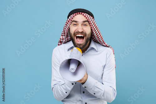 Cheerful bearded arabian muslim man in keffiyeh kafiya ring igal agal casual clothes isolated on pastel blue background. People religious lifestyle concept. Mock up copy space. Scream in megaphone. photo