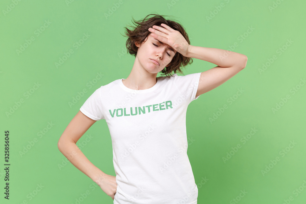 Tired young brunette woman in white volunteer t-shirt isolated on pastel green background. Voluntary free work assistance help charity grace teamwork concept. Put hand on head, keeping eyes closed. Tired young brunette woman in white volunteer t-shirt isolated on pastel green background. Voluntary free work assistance help charity grace teamwork concept. Put hand on head, keeping eyes closed.