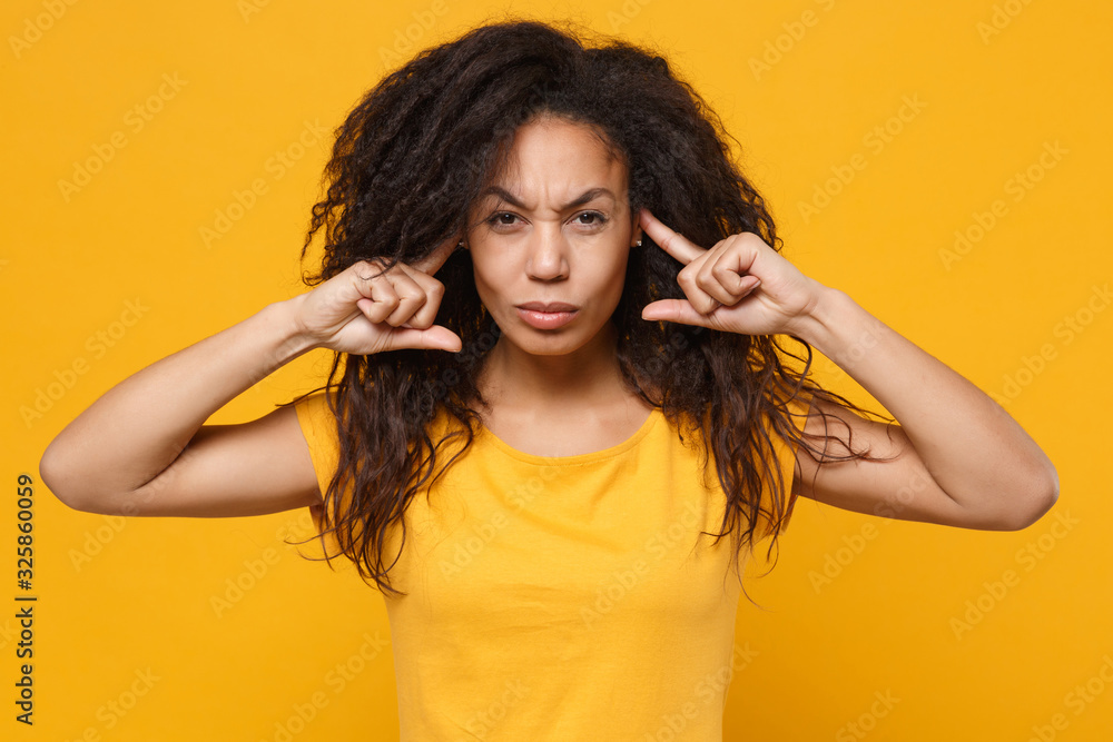 Dissatisfied young african american woman girl in casual t-shirt posing isolated on yellow orange background studio portrait. People lifestyle concept. Mock up copy space. Covering ears with fingers. Dissatisfied young african american woman girl in casual t-shirt posing isolated on yellow orange background studio portrait. People lifestyle concept. Mock up copy space. Covering ears with fingers.