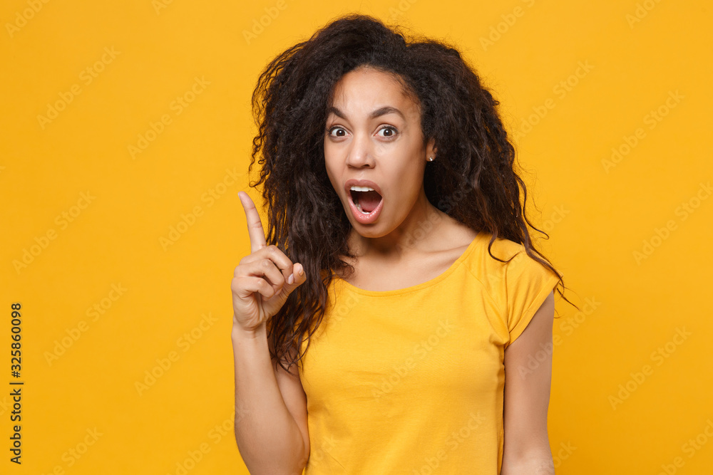Shocked bewildered young african american woman girl in casual t-shirt posing isolated on yellow orange background. People lifestyle concept. Mock up copy space. Pointing index finger up, swearing. Shocked bewildered young african american woman girl in casual t-shirt posing isolated on yellow orange background. People lifestyle concept. Mock up copy space. Pointing index finger up, swearing.