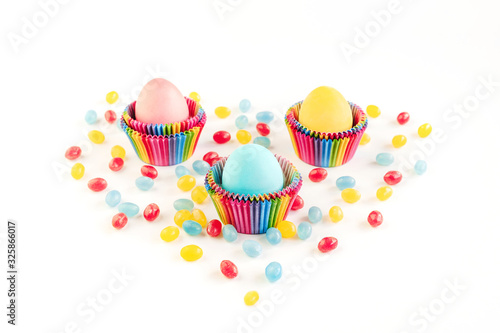 Bright creative Easter decoration of colorful eggs in raibow colored paper forms for cupcakes on white background decorated with jelly candies. Mockup for your design. Copy space.