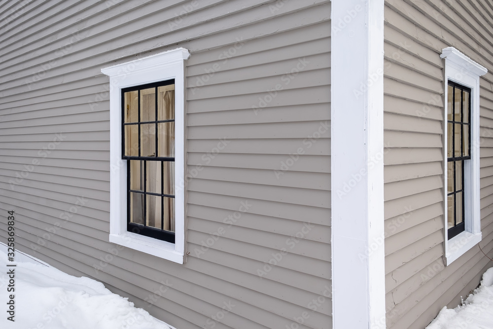 Corner view of a vintage tan-colored house with white trim and black wooden window spacers. The glass in the window is wavy and reflecting the sun. There's snow on the ground and up the building.