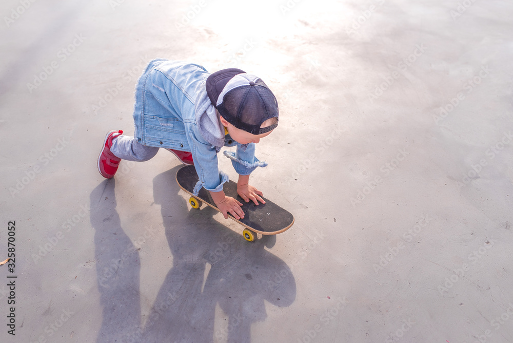 Little boy 3-5 years old, view from the top, in summer on sports ground, learning to ride a skateboard, casual wear, jeans, pants, a baseball cap. Free space for copy text. Little boy 3-5 years old, view from the top, in summer on sports ground, learning to ride a skateboard, casual wear, jeans, pants, a baseball cap. Free space for copy text.