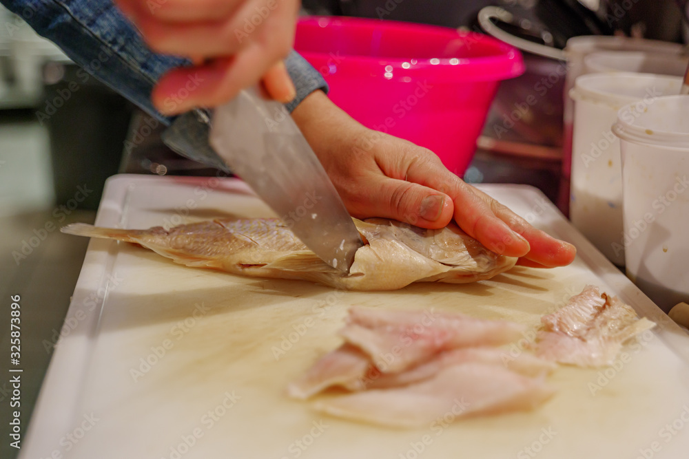 Close up view of Chef'hand use knife for cut and slice red snapper fish on white chopping block and sliced pieces of fish fillet, on countertop in kitchen.