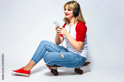 Happy Caucasian Female Blond Girl In Casual Closing Listeining To Music in Headphones On Smartphone. posing on Skateboard Against White. photo