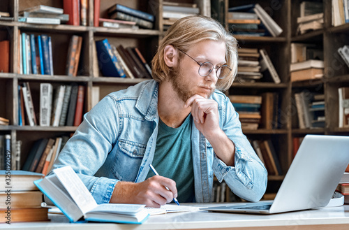 Focused young man student hipster home library desk read look at modern laptop screen make notes has skype course web study online training e learning take test distant electronic education concept.