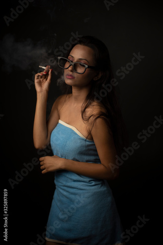 Portrait of Asian/Japanese/Korean/Indian brunette young girl in blue bath towel and spectacles smoking cigarette in black studio copy space background. Indian lifestyle and fashion photography