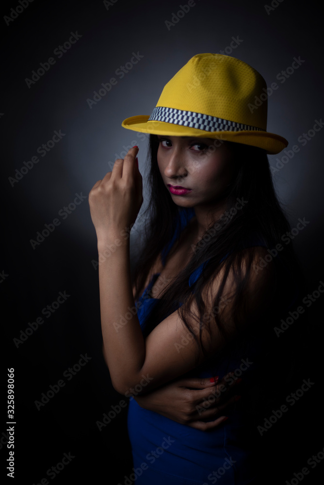 Fashion portrait of an young and attractive Indian Bengali brunette girl in blue western dress and yellow cap in front of a black studio background. Indian fashion portrait and lifestyle. Fashion portrait of an young and attractive Indian Bengali brunette girl in blue western dress and yellow cap in front of a black studio background. Indian fashion portrait and lifestyle.
