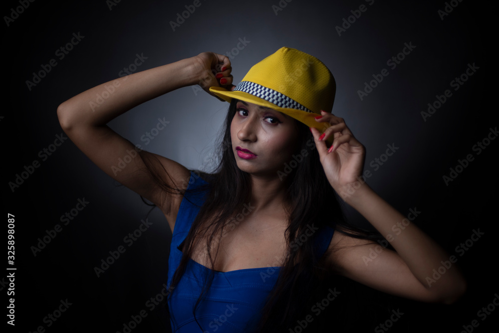 Fashion portrait of an young and attractive Indian Bengali brunette girl in blue western dress and yellow cap in front of a black studio background. Indian fashion portrait and lifestyle. Fashion portrait of an young and attractive Indian Bengali brunette girl in blue western dress and yellow cap in front of a black studio background. Indian fashion portrait and lifestyle.