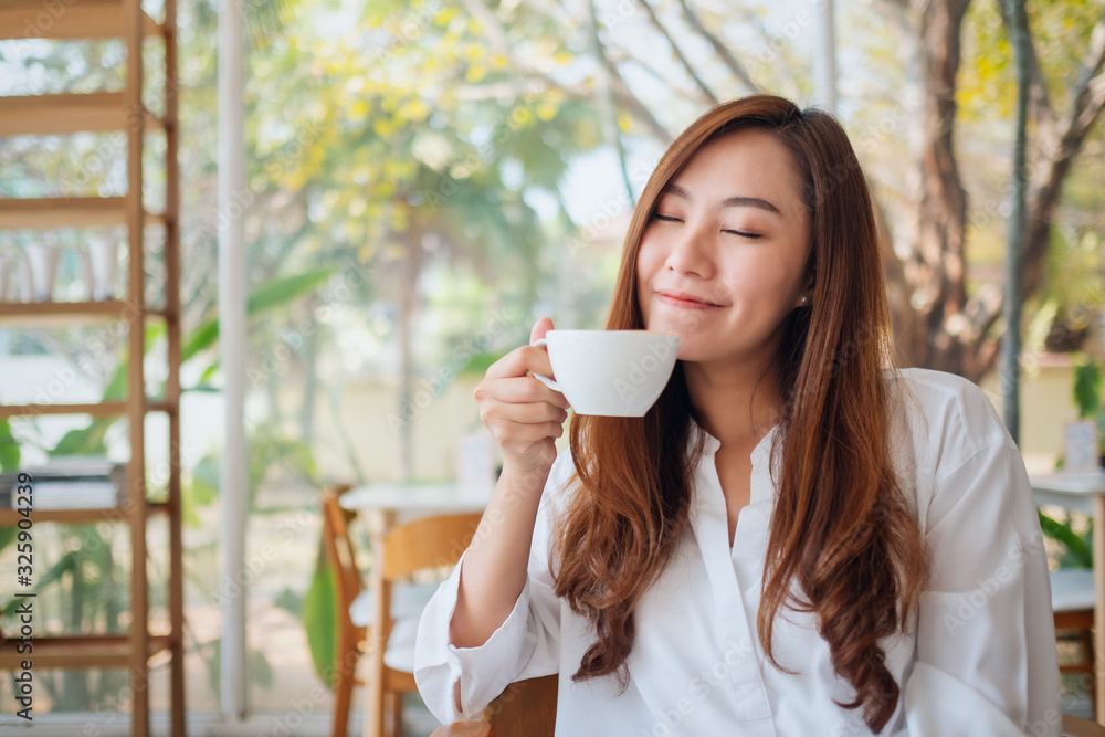 Closeup image of a beautiful asian woman holding and smelling a cup of hot coffee in cafe Closeup image of a beautiful asian woman holding and smelling a cup of hot coffee in cafe