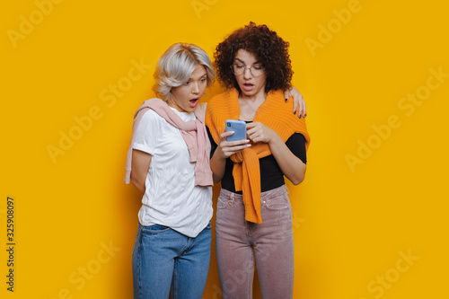 Two curly haired girls feel amazed by something after looking at the phone on a yellow background