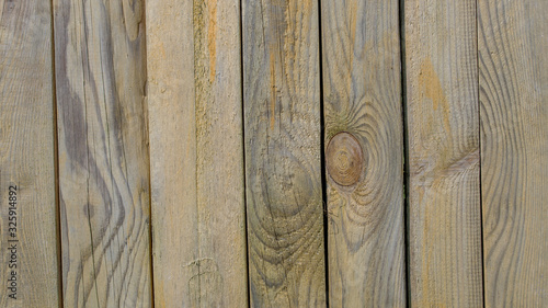 The fence wall is made of vertical wooden treated narrow boards with the texture of knots and streaks of wood, with traces of yellow paint stain on the surface