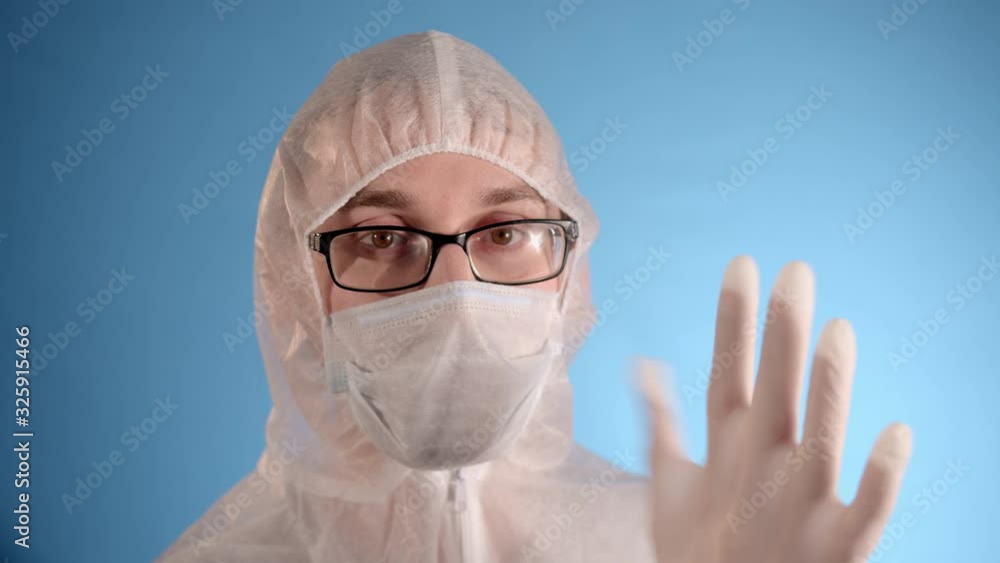 Caucasian man in a white protective suit, glasses and medical mask on a blue background puts rubber white gloves on his hands Caucasian man in a white protective suit, glasses and medical mask on a blue background puts rubber white gloves on his hands