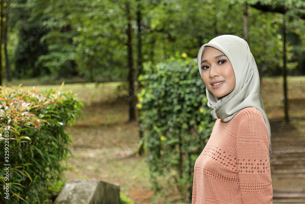 One Asian Malay Muslim woman in headscarf posing smile look forward towards camera confident outdoor green park One Asian Malay Muslim woman in headscarf posing smile look forward towards camera confident outdoor green park