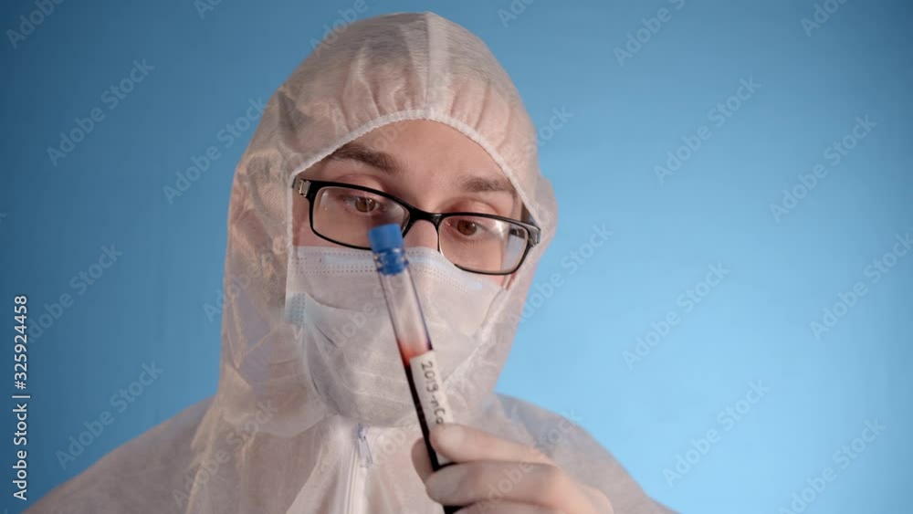 Caucasian man in protective suit, medical mask, glasses, rubber gloves on blue background holds in hands test tube with red liquid and inscription Coronovirus. Then opening it, surprising, sniffing Caucasian man in protective suit, medical mask, glasses, rubber gloves on blue background holds in hands test tube with red liquid and inscription Coronovirus. Then opening it, surprising, sniffing