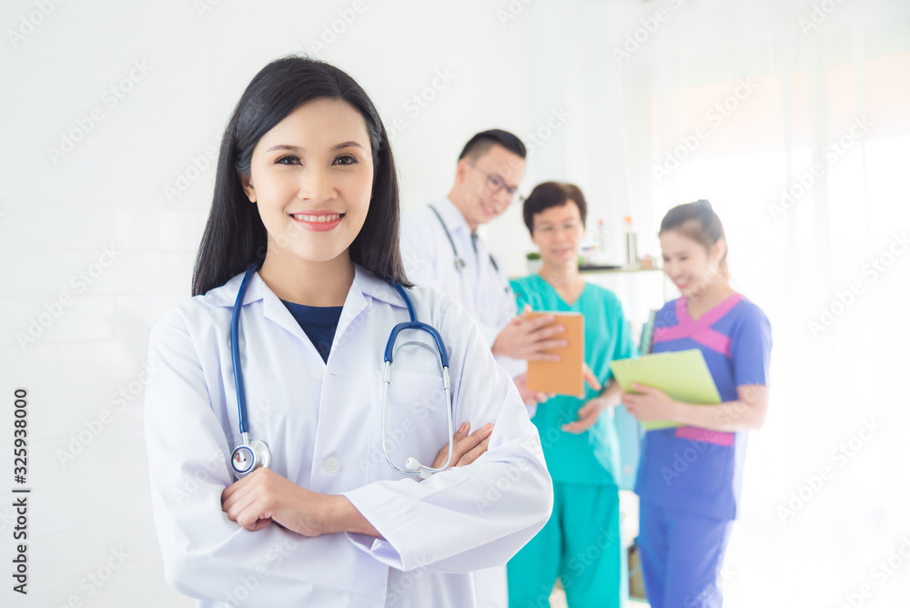 Young asian doctor in white coat smiling at camera with her team standing in background. Young asian doctor in white coat smiling at camera with her team standing in background.