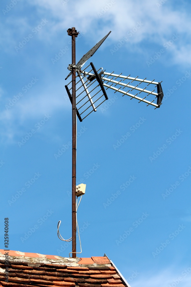 Large TV antenna mounted on top of rusted metal pole on edge of suburban family house roof covered with dilapidated old roof tiles on clear blue sky background Large TV antenna mounted on top of rusted metal pole on edge of suburban family house roof covered with dilapidated old roof tiles on clear blue sky background