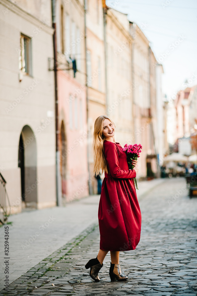 Fototapeta premium Portrait of young stylish beautiful girl in a red dress with bouquet of flowers running on the street, smiling enjoy her weekends. Summer, sunny, trendy, lifestyle.