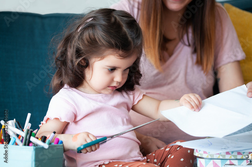 Little curly toddler girl with scissor stay in isolation at home. Education.Portrait of a little Cute baby girl cutting a paper focused on what she is doing.Quality time  at home and happy childhood  photo