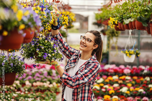 Young smiling female nursery garden worker standing and hanging pot with flowers.