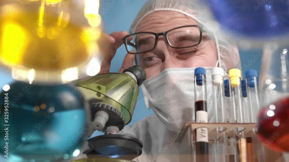 Scientist wearing glasses in medical mask and protective overalls drops red liquid from pipette into beaker. On glass transparent table are vessels and flasks with multi-colored solutions Scientist wearing glasses in medical mask and protective overalls drops red liquid from pipette into beaker. On glass transparent table are vessels and flasks with multi-colored solutions