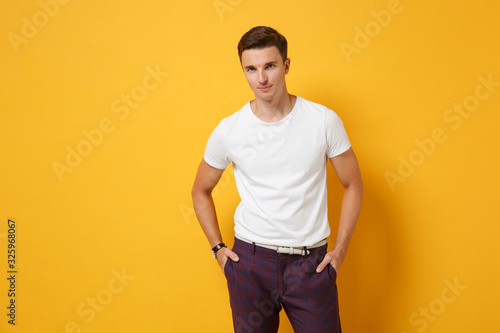 Confident young man guy 20s in white t-shirt posing isolated on yellow orange wall background studio portrait. People sincere emotions, lifestyle concept. Mock up copy space. Holding hands in pockets.