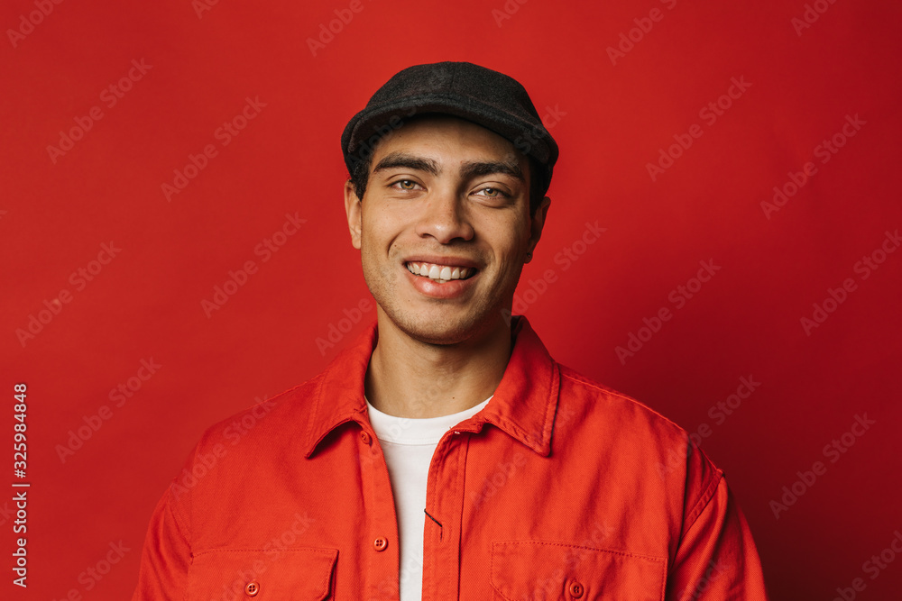 Portrait of cheerful attractive arabian guy look on camera and smile. Hispanic of egyptian young man wear black hat and red shirt. Isolated over red background. Portrait of cheerful attractive arabian guy look on camera and smile. Hispanic of egyptian young man wear black hat and red shirt. Isolated over red background.