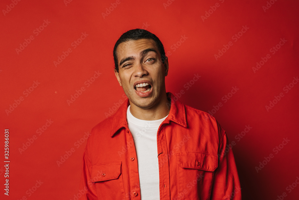 Playful young man having fun and posing on camera alone. Weird facial expressions. Emotional problems. Wear colorful clothes. Isolated over red background. Playful young man having fun and posing on camera alone. Weird facial expressions. Emotional problems. Wear colorful clothes. Isolated over red background.