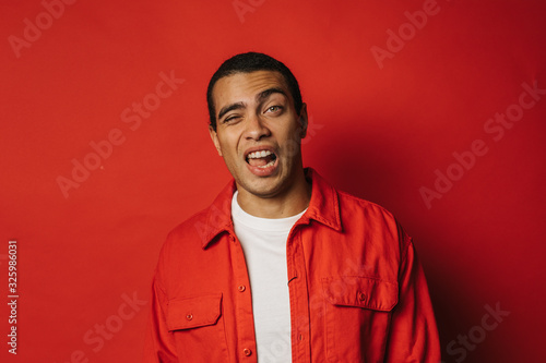 Playful young man having fun and posing on camera alone. Weird facial expressions. Emotional problems. Wear colorful clothes. Isolated over red background.